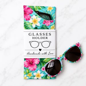 May include: A quilted fabric glasses holder featuring a vibrant tropical print with pink hibiscus and white plumeria flowers on a turquoise background. A white paper band reads GLASSES HOLDER with an icon and Handmade with Love. Matching sunglasses.
