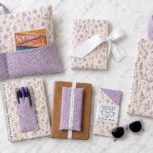 K&ouml;nnte beinhalten: Eine Sammlung von Stoffaccessoires mit hellviolettem Blumenmuster und dunkelviolettem Punktemuster. Die Artikel umfassen ein Buchkissen mit Taschen, einen Notizbuchumschlag mit wei&szlig;em Band, eine Tragetasche, ein Eck-Lesezeichen, einen Stiftehalter, ein Notizbuchband und einen Brillenhalter mit einer schwarzen Sonnenbrille.