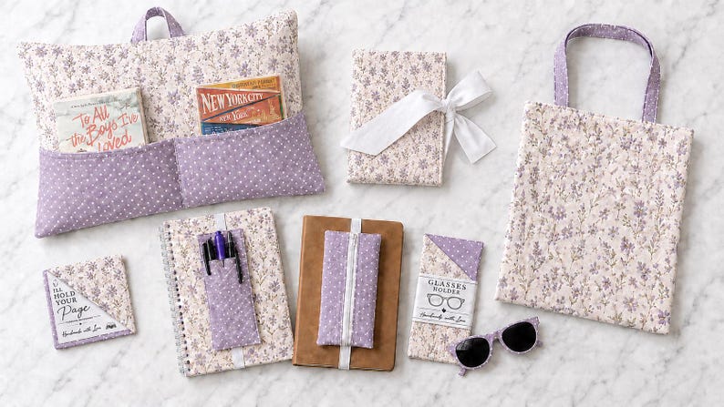 K&ouml;nnte beinhalten: Eine Sammlung von Stoffaccessoires mit hellviolettem Blumenmuster und dunkelviolettem Punktemuster. Die Artikel umfassen ein Buchkissen mit Taschen, einen Notizbuchumschlag mit wei&szlig;em Band, eine Tragetasche, ein Eck-Lesezeichen, einen Stiftehalter, ein Notizbuchband und einen Brillenhalter mit einer schwarzen Sonnenbrille.