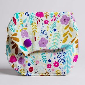 May include: A small, square pouch with a floral pattern. The white fabric is covered in pink, purple, blue, and gold flowers and leaves. A purple button secures the flap closure.