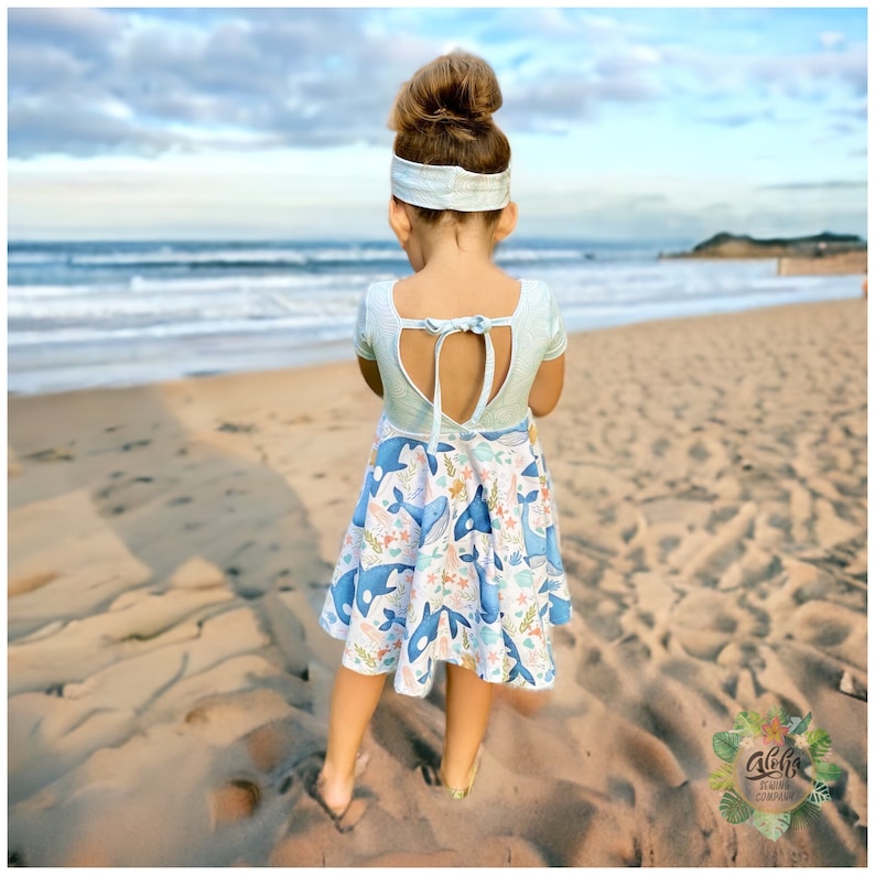 May include: A young girl wearing a light blue dress with a whale print and a light blue headband. The dress has a keyhole back with a tie. The girl is standing on a sandy beach with the ocean in the background.