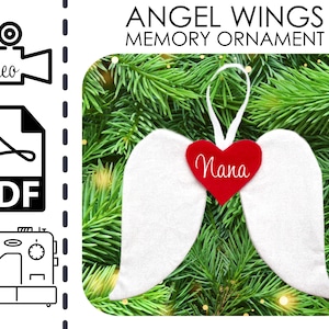 May include: White felt angel wings ornament with a red heart that says "Nana" hanging on a green Christmas tree.