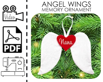 Può includere: Ciondolo a forma di ali d'angelo in feltro bianco con un cuore rosso che dice "Nana" appeso a un albero di Natale verde.