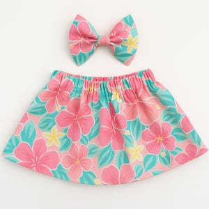 Puede incluir: Un conjunto de falda y lazo a juego con un estampado floral tropical. La falda es de color rosa claro con grandes flores de hibisco rosas, hojas turquesas y detalles amarillos. El lazo tiene el mismo patrón.