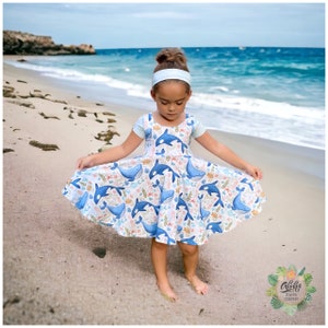 May include: A young girl wearing a blue and white dress with a whale and ocean-themed print. She is standing on a sandy beach with the ocean in the background.