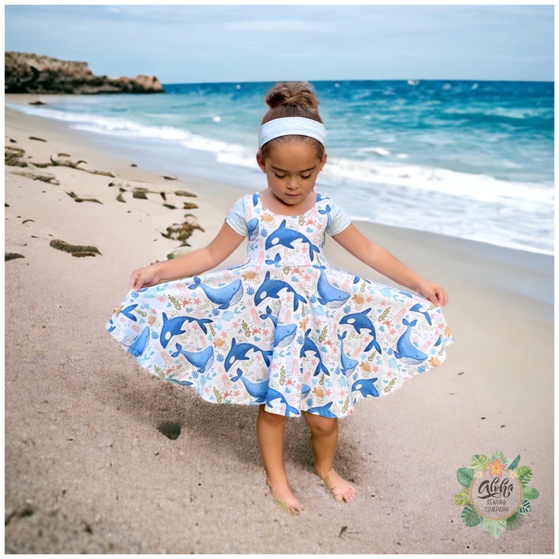 May include: A young girl wearing a blue and white dress with a whale and ocean-themed print. She is standing on a sandy beach with the ocean in the background.
