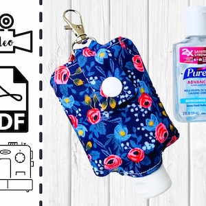 May include: A blue and white floral fabric hand sanitizer holder with a snap closure and a key ring. The holder is attached to a clear plastic hand sanitizer bottle. The bottle is labeled "Purell Advanced Hand Sanitizer" and "Refreshing Gel".
