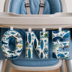 May include: A fabric banner spelling the word ONE, featuring three padded letters hanging from a brown twine string. The letters display blue and white patterns of sharks, surfboards, and palm trees, with small blue pom-pom trim edges.