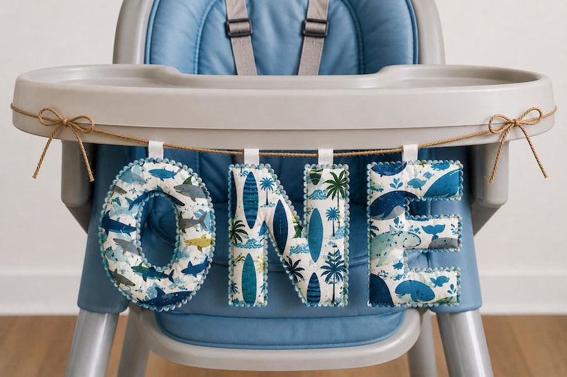May include: A fabric banner spelling the word ONE, featuring three padded letters hanging from a brown twine string. The letters display blue and white patterns of sharks, surfboards, and palm trees, with small blue pom-pom trim edges.