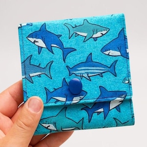 以下が含まれることがあります： 青いサメの模様が入った、小さな正方形の財布。財布は布製で、青いボタン留めが付いています。サメは、ターコイズブルーの背景に、青と白の様々な色合いで描かれています。