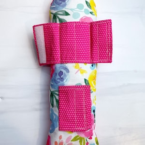 May include: A pink and white floral print pillow with a pink polka dot fabric strap and pocket. The pillow is filled with rice or beans for weight.
