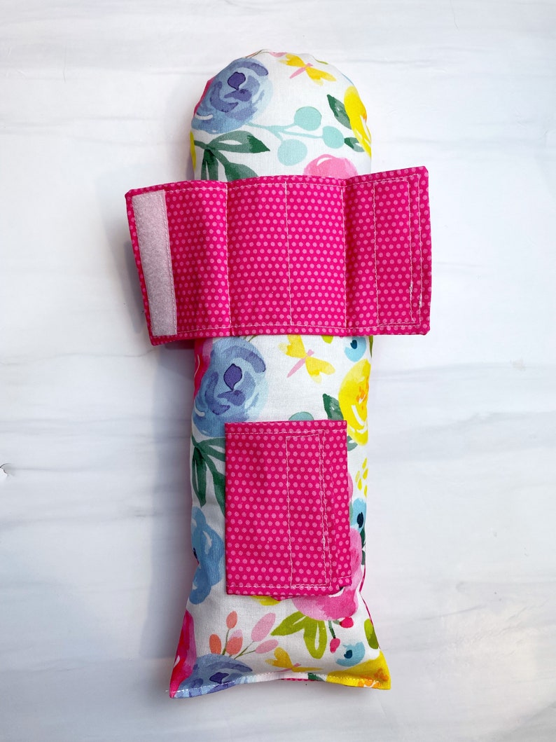 May include: A pink and white floral print pillow with a pink polka dot fabric strap and pocket. The pillow is filled with rice or beans for weight.