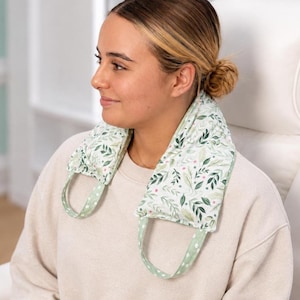 May include: A neck wrap featuring a white fabric base with a botanical pattern of green leaves and small pink accents. The wrap has two integrated fabric handles in a light green colour with a white polka dot pattern, designed to rest over shoulders.