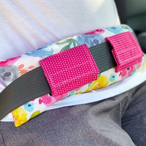 May include: A floral patterned seatbelt cushion with two pink fabric pockets. The cushion is designed to provide comfort and support while wearing a seatbelt.
