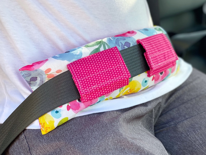 May include: A floral patterned seatbelt cushion with two pink fabric pockets. The cushion is designed to provide comfort and support while wearing a seatbelt.