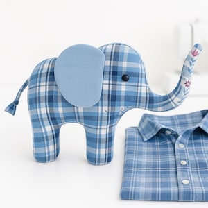 Op de afbeelding: Een blauw geruit olifantenknuffel met een lichtblauw oor en een bloemenmotief op de slurf. Een gevouwen blauw geruit overhemd ligt naast de olifant. De olifant heeft een zwart knoopoog.