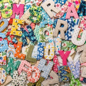 May include: A collection of fabric letters in various patterns and colors, including floral, plaid, and polka dot prints. Each letter features stitched edges. A rectangular fabric label in the bottom right corner reads Aloha Sewing Company.