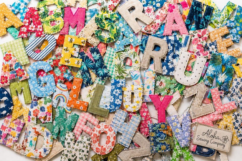 May include: A collection of fabric letters in various patterns and colors, including floral, plaid, and polka dot prints. Each letter features stitched edges. A rectangular fabric label in the bottom right corner reads Aloha Sewing Company.