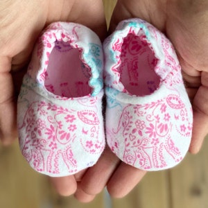May include: A pair of white baby booties with a pink elephant print. The booties have a light blue trim around the top.