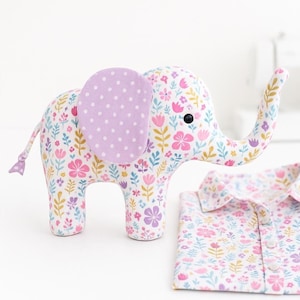 May include: A stuffed elephant toy with a floral pattern in pink, purple, and yellow. The elephant has a lavender ear with white polka dots and a matching tail. A folded floral patterned shirt is next to the elephant.