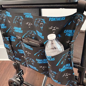 May include: Black fabric walker bag featuring the Carolina Panthers logo in blue. The bag has multiple pockets, one holding a wallet and another a water bottle. The bag is attached to a walker frame.