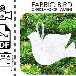 May include: A white fabric bird ornament with a snowflake pattern, hanging on a green pine branch. The ornament is a DIY project with instructions available in PDF and video format.
