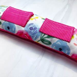 May include: A long, rectangular pillow with a white background and a floral pattern in shades of pink, blue, green, and yellow. The pillow has two pink pockets with white polka dots on the front.