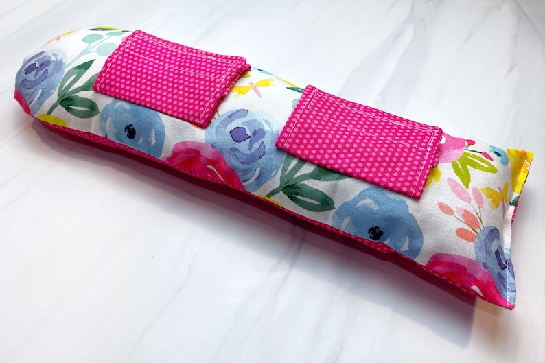 May include: A long, rectangular pillow with a white background and a floral pattern in shades of pink, blue, green, and yellow. The pillow has two pink pockets with white polka dots on the front.