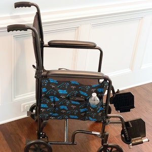 May include: A black and silver transport wheelchair with a Carolina Panthers logo on the storage bag. The wheelchair has a black seat, armrests, and footrests. A water bottle is in the side pocket.