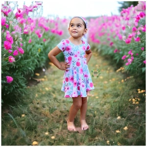 May include: A young girl wearing a floral print dress and a white headband stands in a field of pink flowers. She is looking up and smiling.
