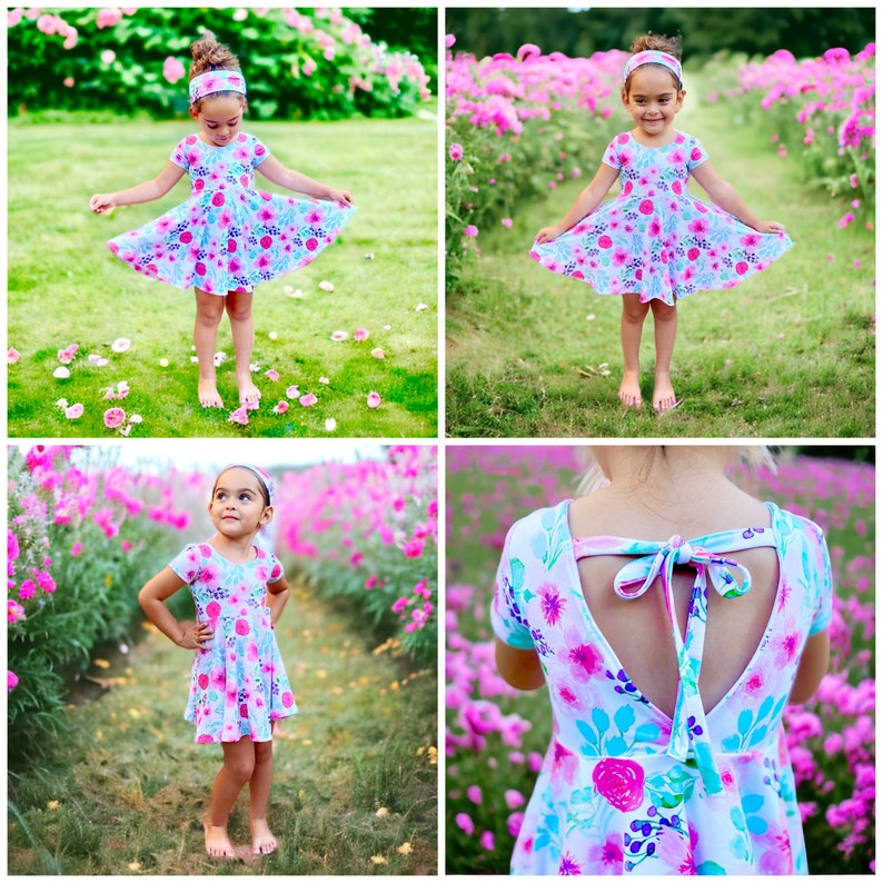 May include: A young girl wearing a light blue and pink floral print dress with a matching headband. The dress has a low back with a bow tie detail. The girl is standing in a field of pink flowers.