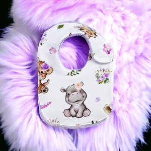 May include: White baby bib with a purple floral and animal print. The bib features a hippo, a giraffe, and a lion. The bib has a white velcro closure.