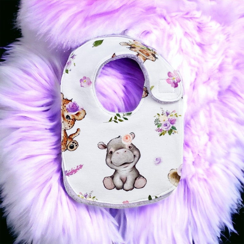 May include: White baby bib with a purple floral and animal print. The bib features a hippo, a giraffe, and a lion. The bib has a white velcro closure.