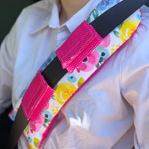 May include: A pink and white floral patterned seatbelt shoulder pad with a black strap and two pink pockets. The pad is designed to provide comfort and support while wearing a seatbelt.