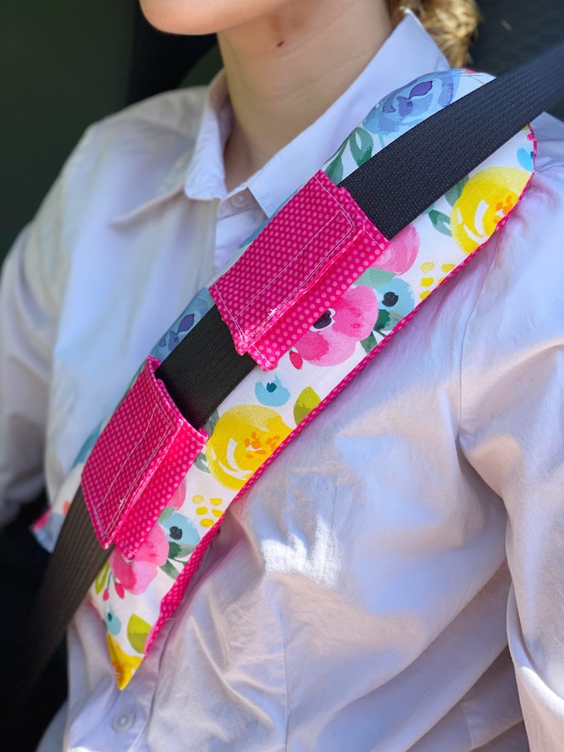 May include: A pink and white floral patterned seatbelt shoulder pad with a black strap and two pink pockets. The pad is designed to provide comfort and support while wearing a seatbelt.