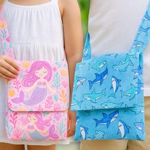 Könnte beinhalten: Zwei Umhängetaschen. Eine ist rosa mit einem Meerjungfrauen-Design, mit einer Meerjungfrau mit rosa Haaren und einem lila Schwanz, umgeben von Muscheln und Seetang. Die andere ist blau mit einem Hai-Muster. Beide Taschen haben passende Träger.