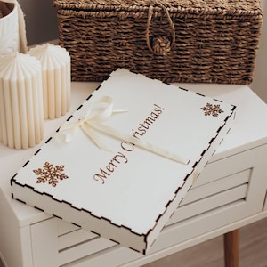 Könnte beinhalten: Eine rechteckige weiße Holzschachtel mit Klappdeckel, verziert mit dem braunen Schriftzug Merry Christmas, braunen Schneeflockenmotiven und einer cremefarbenen Schleife, platziert auf einem weißen Nachttisch mit hölzernen Beinen.