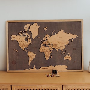 May include: A wooden world map with a dark grey background and a light brown frame. The map is detailed with country names and is displayed on a light brown cabinet. A white lamp with a gold interior and candles are also visible.