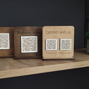 Peut inclure: Trois panneaux en bois avec des codes QR de différentes nuances de brun. Le panneau de droite indique "Connectez-vous avec nous" avec les logos Facebook et Instagram. Un petit pot carré blanc avec de la mousse verte est à droite.