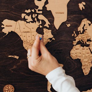 May include: A wooden world map with a dark brown finish, featuring individual country outlines in a lighter wood tone. A hand with a blue push pin is placed on the map. A compass rose is in the lower left corner.