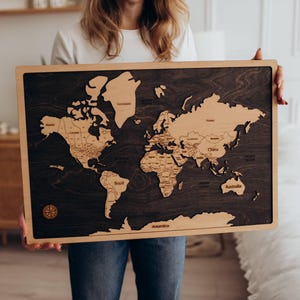 May include: Wooden world map with a dark stained background and light wood continents. The map is framed in a light wood border and includes country names. A compass rose is in the lower left corner. The map is being held by a person.