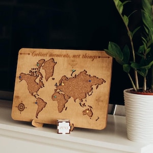 May include: A wooden world map with push pins, a white dresser with drawers, and two green plants in white pots. The map has the text "Collect moments, not things" at the top.