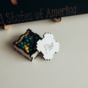 Op de afbeelding: Een klein vierkant houten doosje met een wit deksel waarop het woord Pins in cursief schrift staat. Het doosje bevat diverse punaises met een ronde kop in de kleuren blauwgroen en geel. Het heeft in elkaar grijpende randen en een zwarte binnenkant.