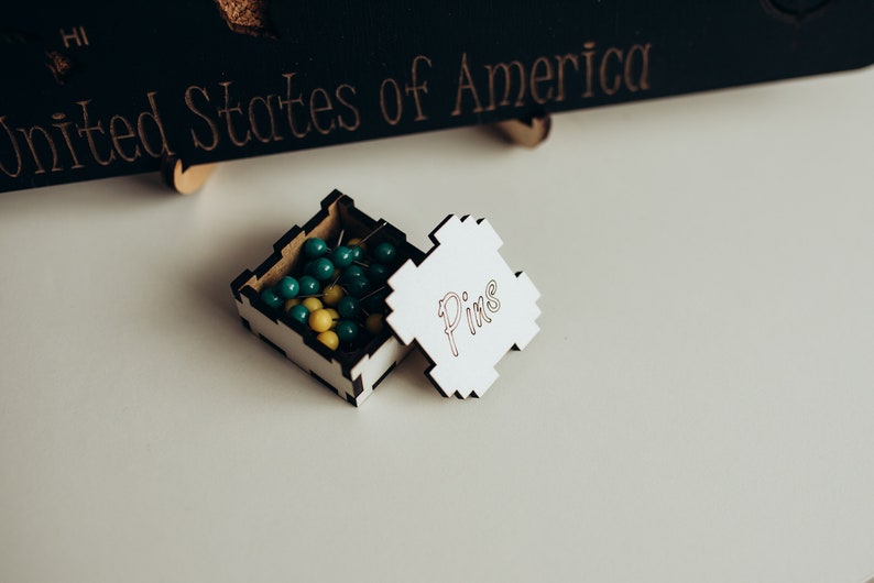 Op de afbeelding: Een klein vierkant houten doosje met een wit deksel waarop het woord Pins in cursief schrift staat. Het doosje bevat diverse punaises met een ronde kop in de kleuren blauwgroen en geel. Het heeft in elkaar grijpende randen en een zwarte binnenkant.
