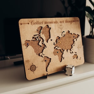 Puede incluir: Un tablero de corcho rectangular con forma de mapa del mundo en madera que incluye el texto grabado con láser "Collect moments, not things" en la parte superior. El tablero presenta un diseño de rosa de los vientos y varias chinchetas.