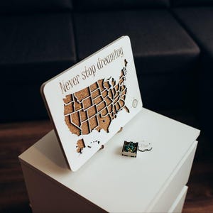 Op de afbeelding: Een wit rechthoekig bord met een kurken uitsparing van de kaart van de Verenigde Staten met staatsgrenzen. De tekst Never stop dreaming is in het zwart boven de kaart gedrukt. Een klein houten doosje met gekleurde spelden staat ernaast.