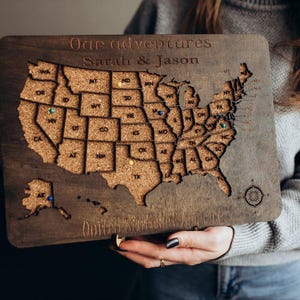 Op de afbeelding: Een rechthoekige houten kaart van de Verenigde Staten met een kurken inleg voor elke staat. Het bord is gegraveerd met de tekst Our adventures, Sarah & Jason en United States of America. Kleine gekleurde punaises markeren specifieke locaties.
