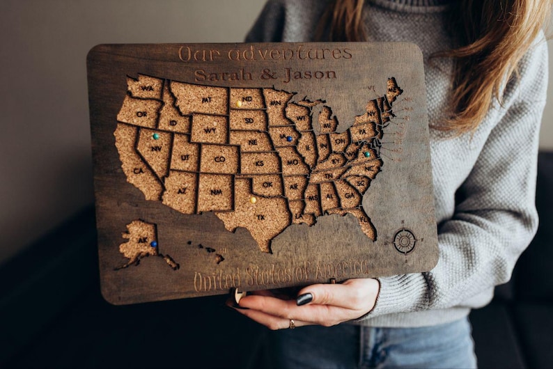 Op de afbeelding: Een rechthoekige houten kaart van de Verenigde Staten met een kurken inleg voor elke staat. Het bord is gegraveerd met de tekst Our adventures, Sarah & Jason en United States of America. Kleine gekleurde punaises markeren specifieke locaties.