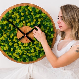 May include: A circular wall clock featuring a wooden frame and a face covered in moss. The clock hands are black, with a red second hand. The clock's diameter is approximately 30 cm.