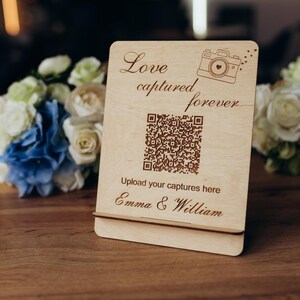 Puede incluir: Un letrero de madera clara con soporte que presenta el texto grabado Love captured forever, un icono de cámara, un código QR, el texto Upload your captures here y los nombres Emma & William, colocado en posición vertical sobre una superficie de madera.
